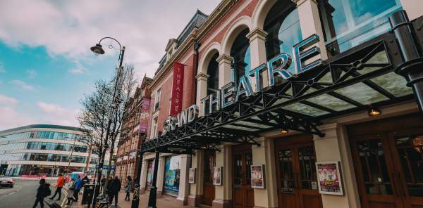 Exterior of the Grand Theatre in Wolverhampton. Large letters GRAND THEATRE on top of a metal canopy covering the 4 double door entrance to the theatre on a busy street.