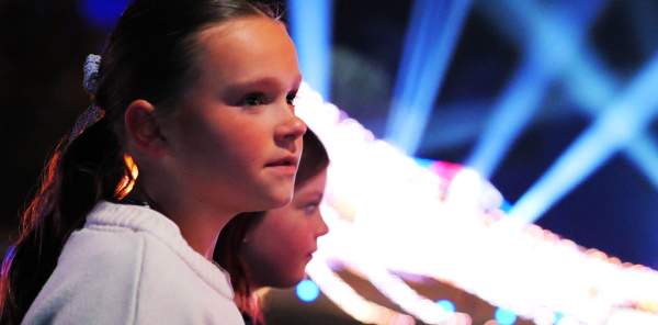 Two girls looking at a Christmas Light show at Warwick Castle. The girl in the foreground has brown hair in a ponytail and is wearing a white jumper. There are spotlights and fairylights blurred in the background, in the distance, behind them both.