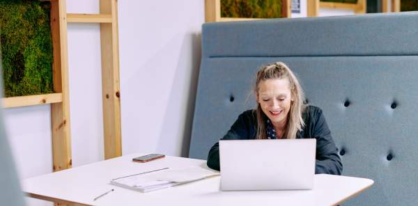 A woman sits at a booth in a workspace using a laptop