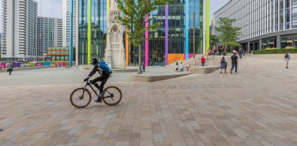 A cyclist crosses in front of the fountain in Paradise, Birmingham, with city dressing for the 2022 Commonwealth Games in the background