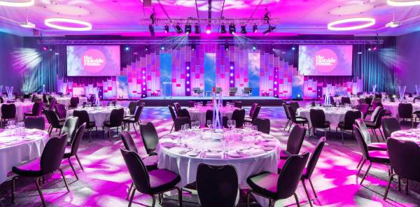 The inside of a large venue, with tables and chairs waiting for guests. Bright purple and pink lighting floods the room, and two projections on either side of the stage read "The Eastside Rooms"