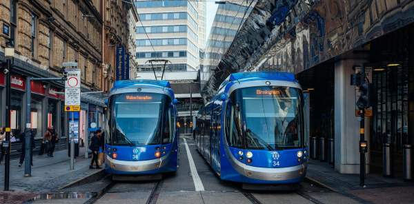 Two blue trams, parked side by side in a street, let passengers embark.