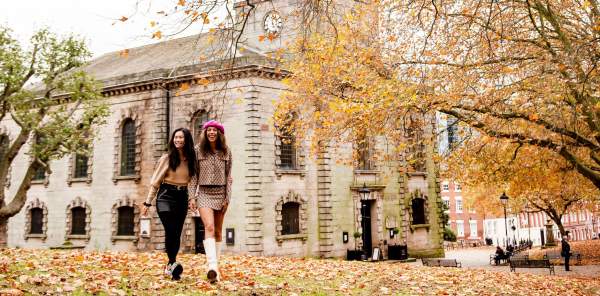 Two friends walking through a park in autumn with fallen orange and red leaves with St Phillip's Cathedral in the background