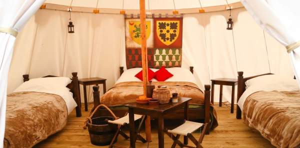 A glamping bedroom at Warwick Castle featuring three bed with fur throws, a small dining table with two chairs and two bed side tables