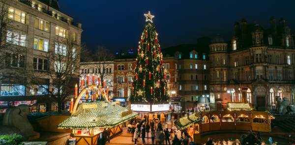 A tall Christmas tree stands in a square in Birmingham at night, the lights are lit and people are gathered round its base at Christmas market stalls