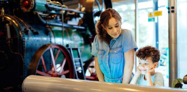 A brother and sister look at a display together at the ThinkTank museum with a steam engine in the background
