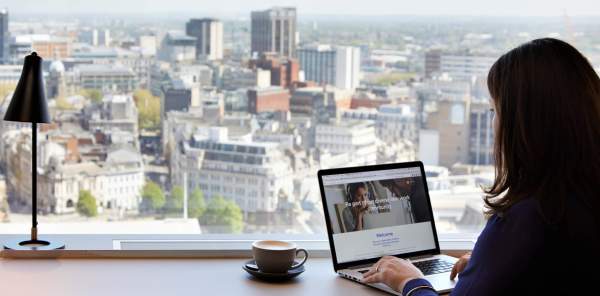 Girl on laptop with a hot drink and view of Birmingham's skyline out of the window