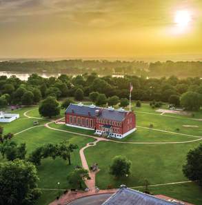 The sun is shining behind clouds above the National Historic Site in Fort Smith, Arkansas with the Arkansas River flowing behind it.
