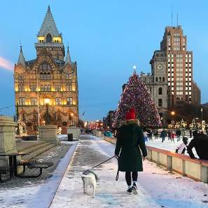 a person walking a dog towards a historic building on a snowy path with a holiday decorated ice skating rink to the side
