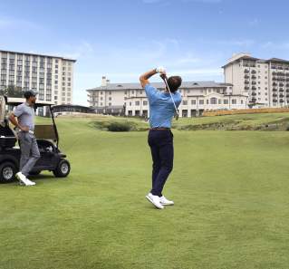 Two men on a golf course, one mid swing, and one leaning on a golf cart.
