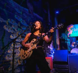 Woman standing on a stage painted with an irredescent fish backdrop, playing the electric guitar and singing into a microphone.