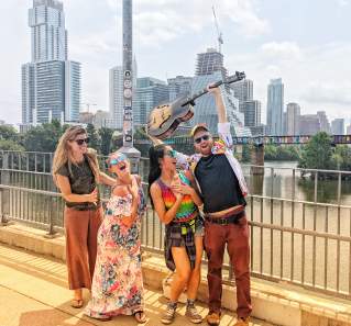 Four people posing on a bridge in front of Austin downtown skyline. Left to Right: woman grins at women beside her, woman places hand over heart and looks towards camera, woman places hands over her heart, man raises a guitar over his head