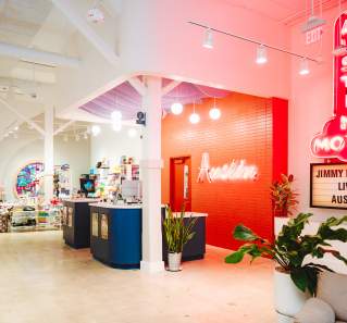 Interior of the Austin Visitor Center showing a welcome desk with brightly colored wall and "Austin" neon sign behind, and a neon Austin Motel sign with marquee reading "Jimmy Kimmel Live! Austin".