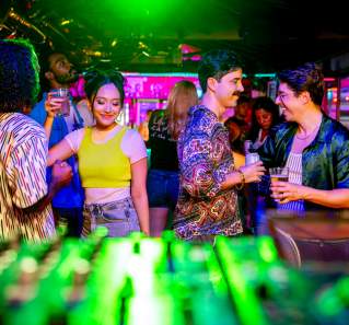 Couples dancing with cocktails in hand in front of a DJ booth with neon lights in an intimate space.