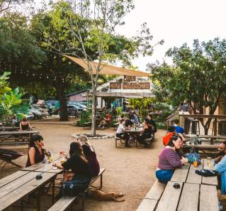 Groups of people sitting at picnic tables in a courtyard surrounded by greenery and a covered patio in the background with the words "ALL ARE WELCOME" posted to the front of the structure.