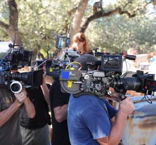 Film crew holding large cameras on a movie set next to an old rusty truck.