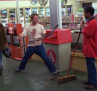 Suburbia screengrab, showing a man in a red shirt sweeping the ground while another man stands in front of him with a hockey stick in his hand. A group of people are walking past them and laughing