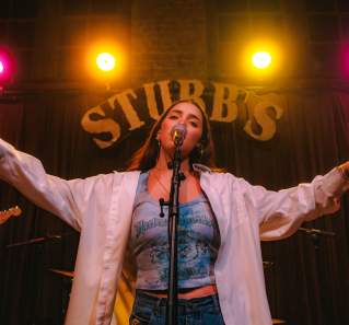 Woman standing in front of a microphone, singing with arms out, a large sign saying "STUBB'S" with pink and yellow neon lights is above her head.