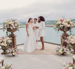 Two women at a wedding altar overlooking Lake LBJ, holding hands with an officiant in the background.