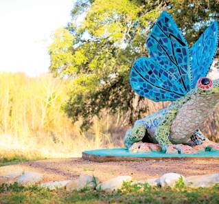 Colorful mosaic sculpture of a frog with a large blue frill, set against a blurred natural background with trees and grass.