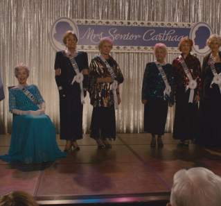 Bernie screengrab showing Bernie on stage as the announcer at the Mrs. Senior Carthage Contest. 8 contestants are on the stage wearing white sashes