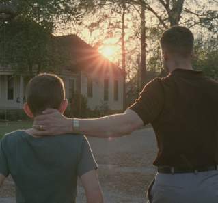 Tree of Life Screengrab, showing a father placing his hand on his son's neck as they walk towards a house. The sunset is visible in front of them