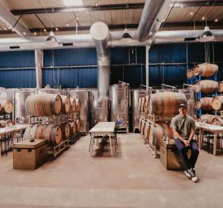 Image of the warehouse at Austin Winery with rows of barrels and the owner standing nearby smiling at the camera.