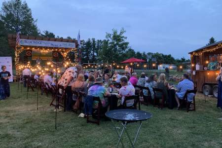 Outdoor gathering in Loudoun, with people seated at long tables under string lights, enjoying food and music. A stage is visible in the background.
