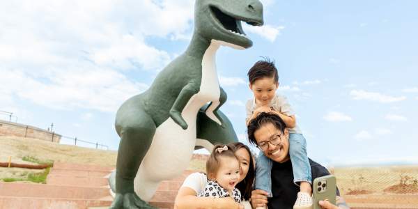 A happy family takes a selfie in front of a large green and white dinosaur statue on a sunny day, with smiles and playful expressions.