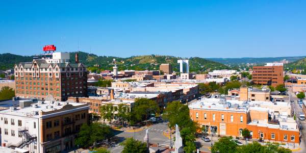 Aerial view of downtown Rapid City featuring historic brick buildings, tree-lined streets, and the Hotel Alex Johnson sign, with green Black Hills in the background under a clear blue sky.