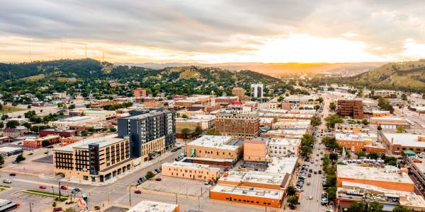 Aerial view of downtown Rapid City at sunset, with downtown streets lined by low-rise buildings, cars, and parking lots. Rolling green hills surround the city, while dramatic clouds stretch across the sky and golden sunlight glows on the horizon.