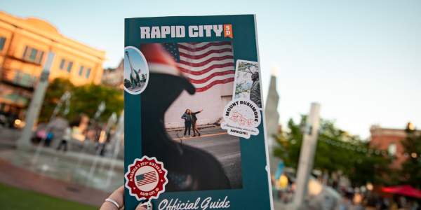 The cover of the Visit Rapid City guide is held up by a woman against the backdrop of the fountains at Main Street Square.