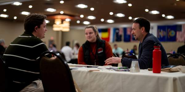 A man and woman sit at a table-cloth draped table across from a man in a striped shirt in conversation.