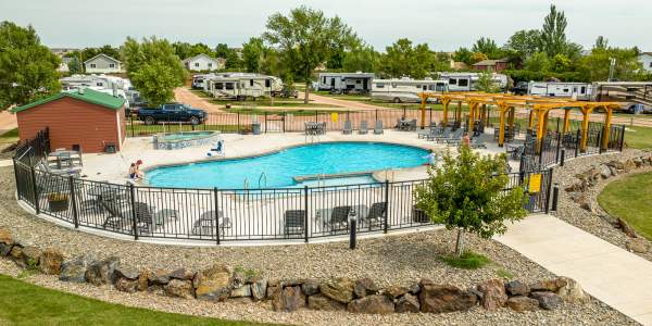 Outdoor pool with blue water heavily distorted by a digital glitch, partially revealing a drained pool basin with tiled walls, concrete edges, and a rocky border, with RVs and trees in the background under a clear sky.