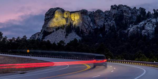 Mount Rushmore, lit up by yellow floodlights at dusk, sits in the background as a vehicle traveling down the highway is trailed by a long line of red taillights, created by a long-exposure photo.