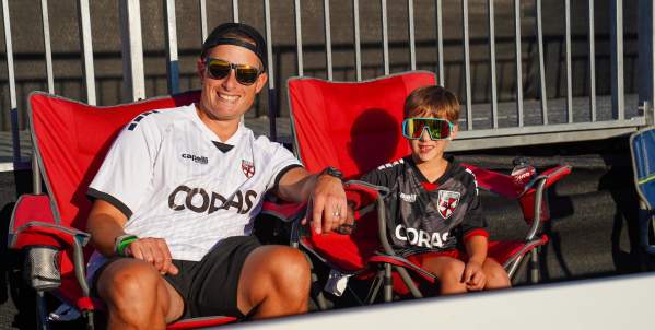 An adult and child wearing Loudoun United FC jerseys sit in red field-level chairs at Segra Field in Leesburg, Virginia