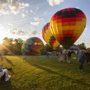 Hot Air Balloons Sit in the Distance as Visitors Watch in Grass and Sun Begins to Set