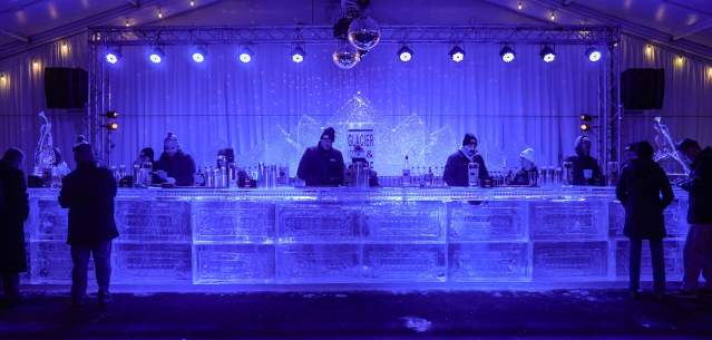 Several bartenders serving drinks at a bar made of ice under deep blue lights.