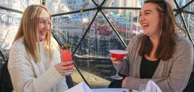 Two women holding drinks and laughing while dining inside an igloo at The Queensbury Hotel
