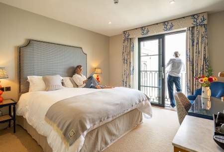 A man and a woman in a hotel room. Woman reading on the bed, man looking out of the window.