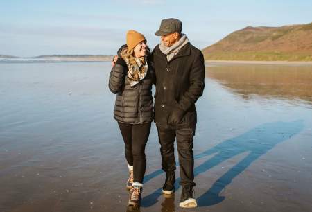 A man and a woman in winter clothes walking arm in arm on a beach.