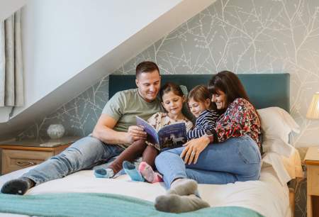 A man, woman and two girls reading a story book on a bed.