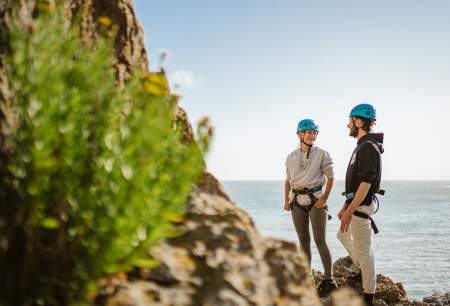 A man and a woman wearing climbing gear smiling at each other next to a cliff.