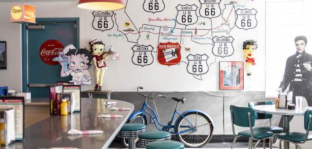 Interior of a retro-themed diner featuring Route 66 and Coca-Cola decorations, teal booths, and a vintage bicycle displayed against a colorful mural wall.