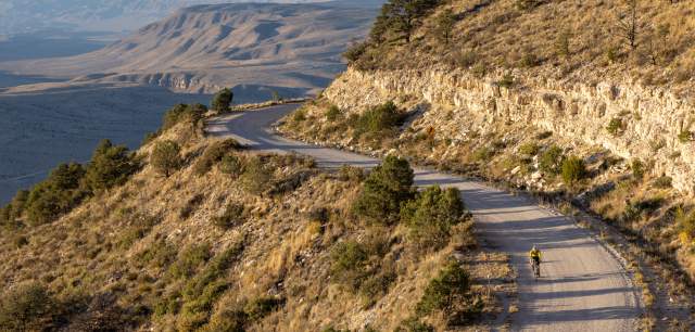 A person cycling on a winding mountain road at sunset, with expansive views of layered hills in the background.