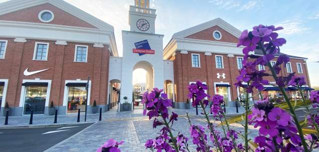 The entrance to the McArthurGlen West Midlands shopping village, on a sunny day, with colourful purple flowers in the foreground.