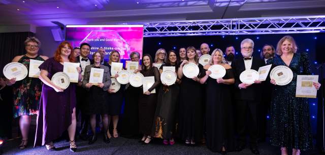 A group shot of the winners of the 2025 Staffordshire & Stoke-on-Trent Tourism Awards. There are about 20 people on a stage, wearing glamorous dresses and tuxedos, and holding trophies and certificates.