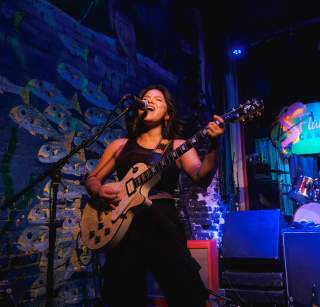 Woman standing on a stage painted with an irredescent fish backdrop, playing the electric guitar and singing into a microphone.