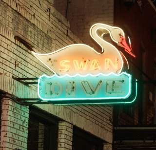 Exterior of a brick bar at night with a neon sign of a swan reading Swan Dive in white and blue lights.