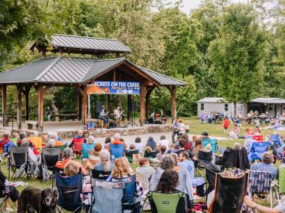 A large crowd of people sit in portable camp chairs on a grassy hill, watching two musicians perform acoustic music under a large wooden pavilion with a metal roof. A banner on the pavilion reads, "Concerts on the Creek - Fridays 7-9 PM." A black dog looks toward the camera in the foreground.